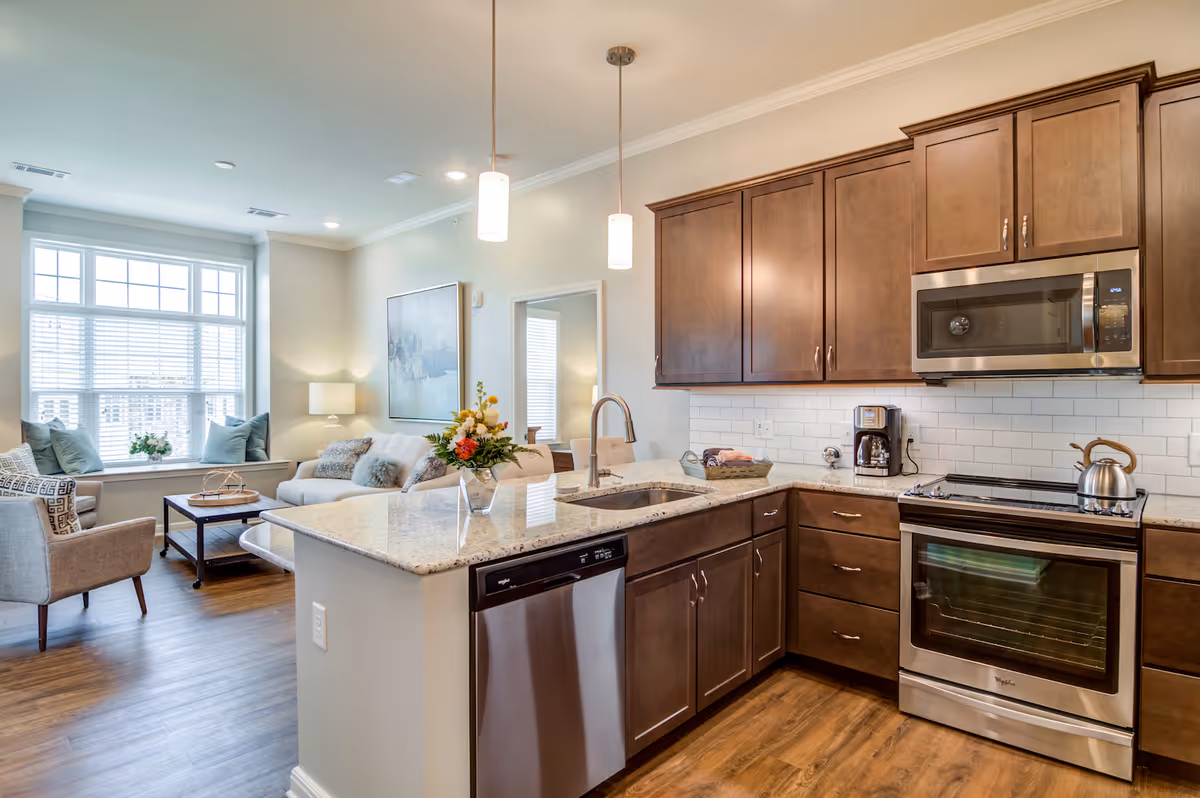 Modern kitchen with wooden cabinets, stainless steel appliances including a microwave, oven, and dishwasher. The kitchen has a granite countertop island with a sink and two pendant lights hanging above. In the background, there is a living room area with a large window, a sofa, armchairs, a coffee table, and decorative cushions.
