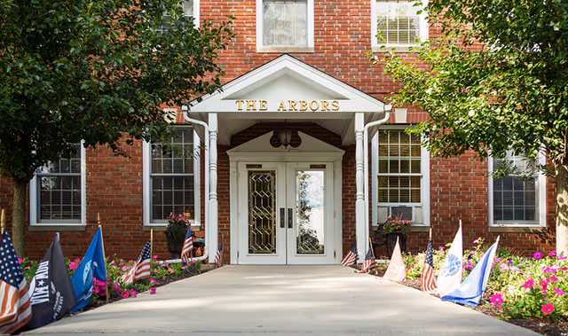 Entrance to a red brick building with a white portico labeled 'THE ARBORS'. The walkway leading to the double glass doors is lined with small American flags and other flags, with green trees and colorful flowers on either side.