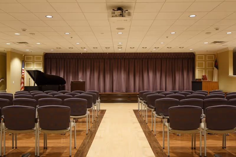 An empty auditorium or meeting room with rows of chairs facing a stage with a closed curtain. On the stage, there is a grand piano on the left and a podium in the center. The room has a drop ceiling with recessed lighting and wood flooring with a light-colored central aisle.