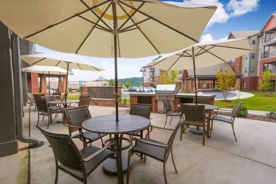 Outdoor patio area with round tables, chairs, and large beige umbrellas. In the background, there is a built-in grill and a landscaped area with grass, trees, and a multi-story residential building under a partly cloudy sky.