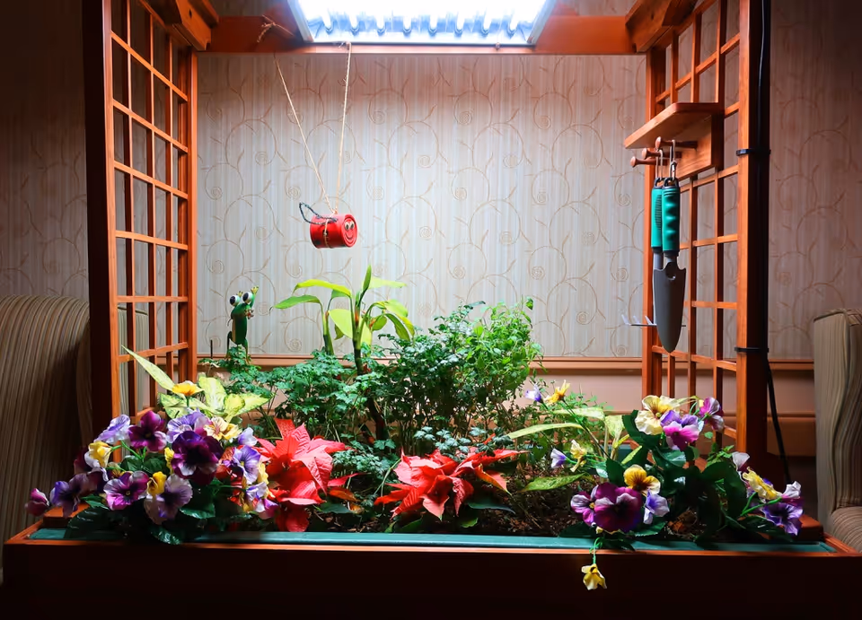 Indoor garden with various colorful flowers and green plants under a skylight, surrounded by wooden lattice panels and gardening tools hanging on the right side.