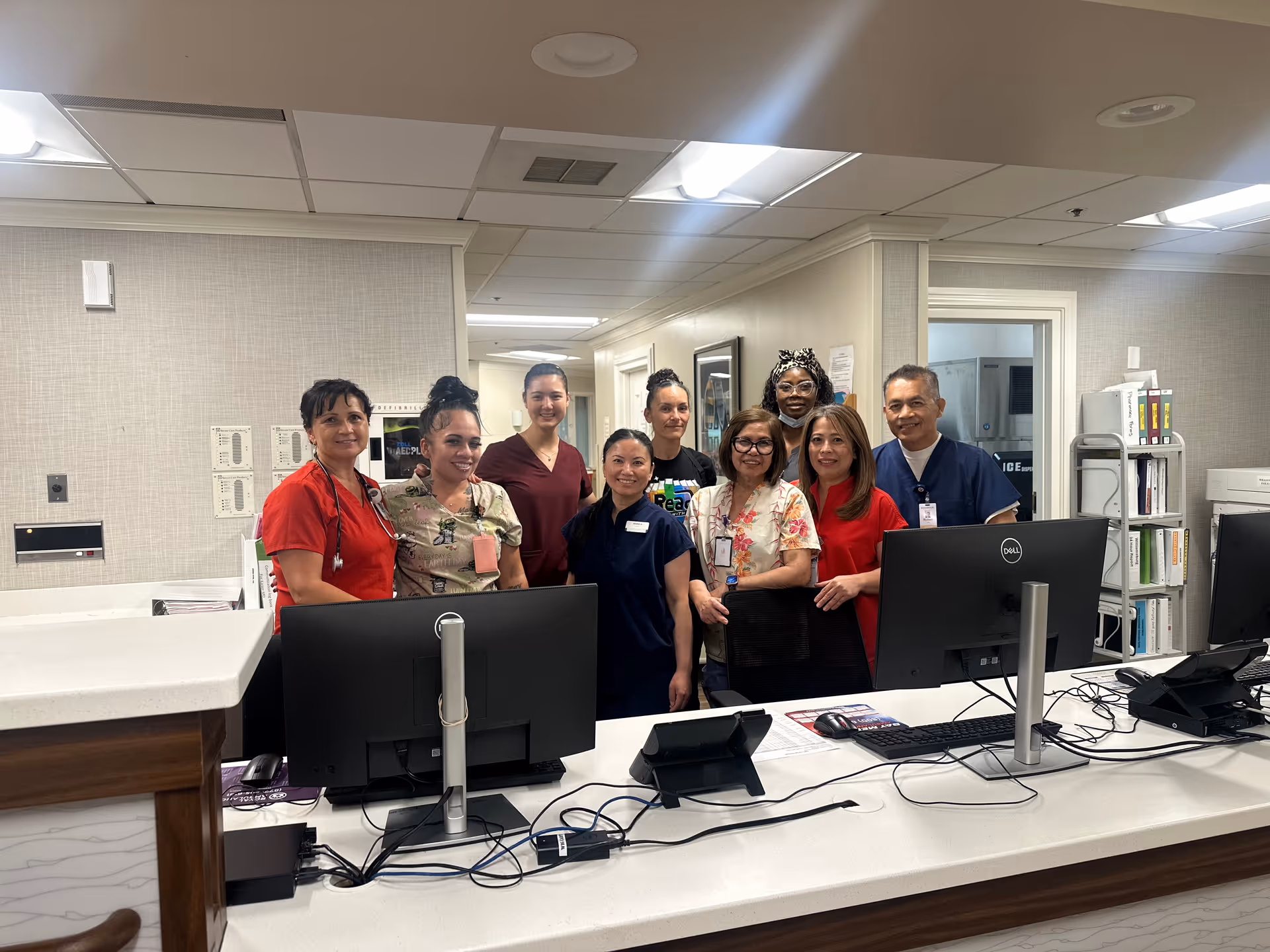A group of nine healthcare workers standing behind a reception desk in a medical or senior care facility. They are smiling and wearing various colored scrubs and ID badges. The desk has computer monitors, keyboards, and office supplies. The background shows a hallway and office doors.