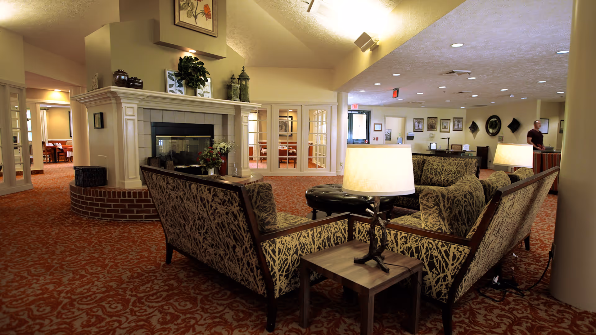 A cozy senior living facility common area with patterned carpet, two upholstered sofas facing each other around a small wooden table with a lamp, and a fireplace with decorative items on the mantle. In the background, there are glass doors leading to a dining area and a person standing near a reception desk.
