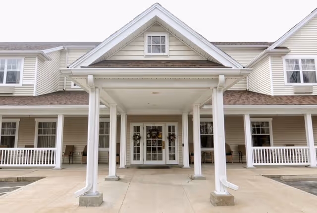 Front entrance of a senior living facility with a covered porch supported by white columns, beige siding, multiple windows, and double glass doors decorated with wreaths.