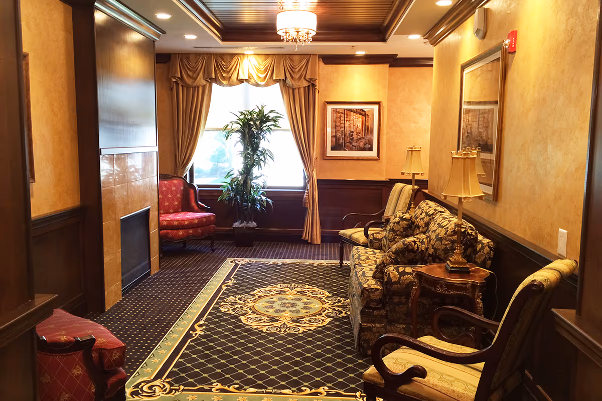A cozy living room with classic decor featuring a patterned carpet, floral upholstered sofa, wooden chairs with cushions, a fireplace, a large window with heavy drapes, a potted plant, framed artwork on the walls, and table lamps on side tables.
