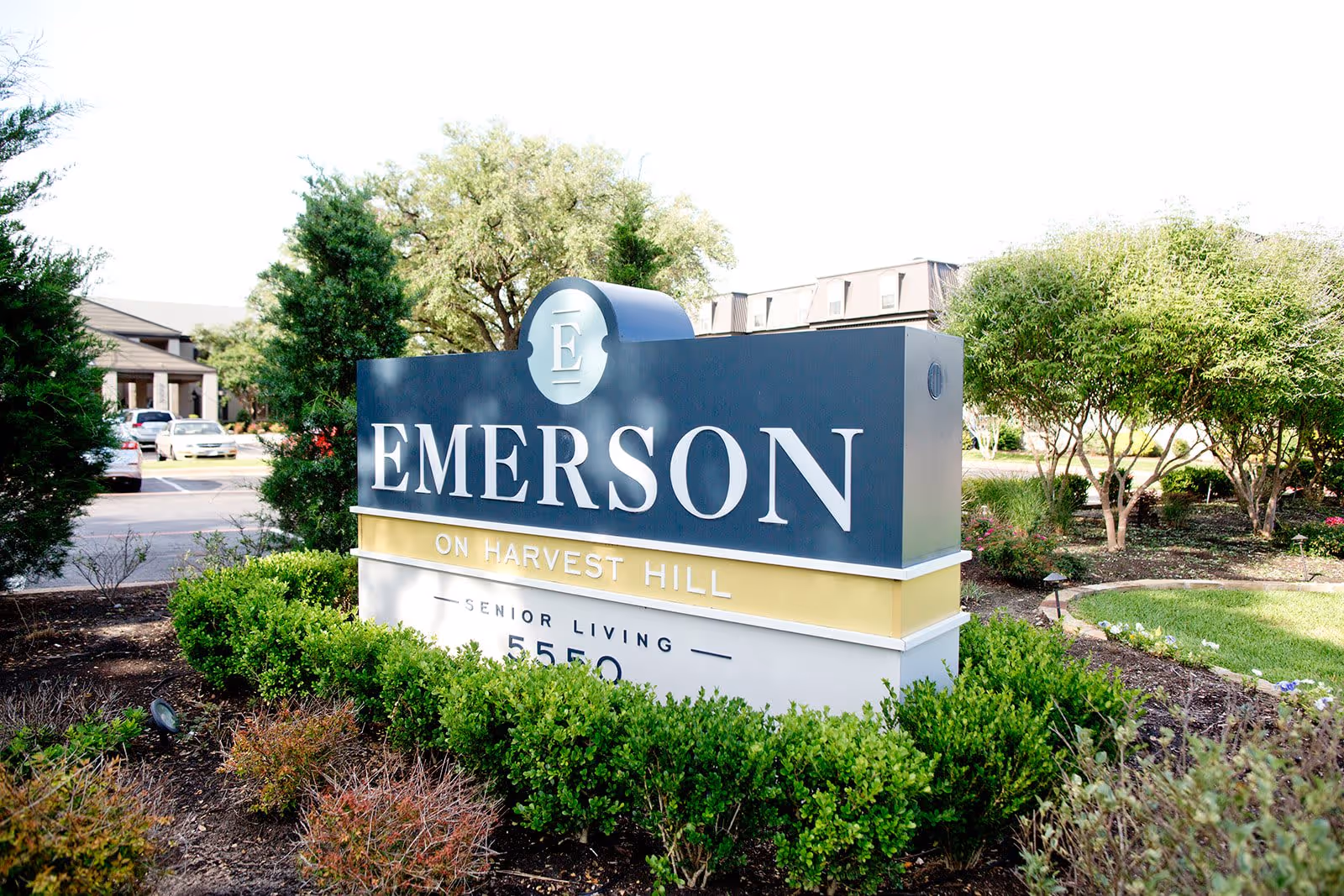 Outdoor view of a large sign for Emerson on Harvest Hill senior living facility surrounded by green bushes and trees with part of the building and parked cars visible in the background.