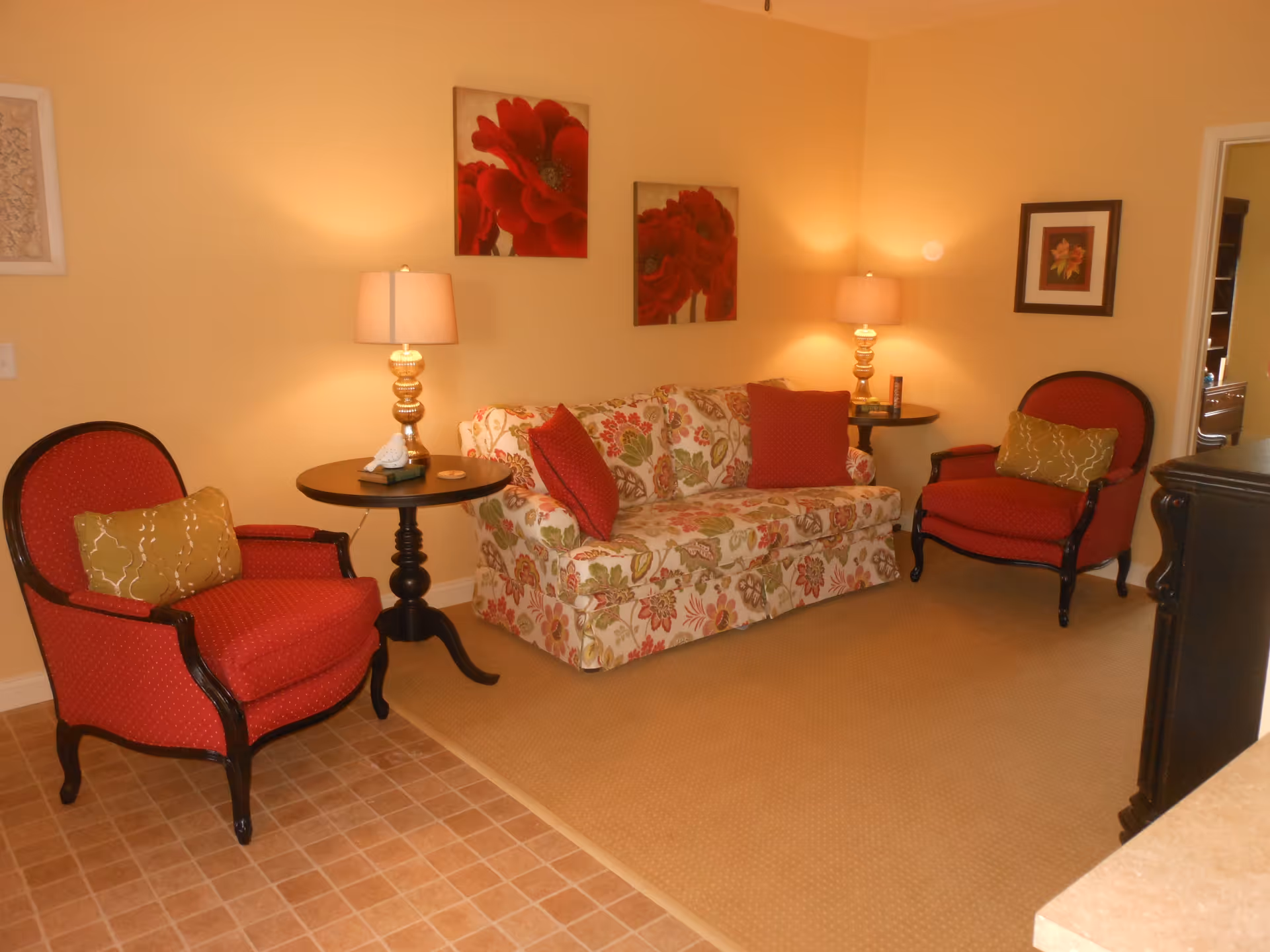 Cozy living room with a floral sofa, two red armchairs, side tables with lamps, and floral wall art.