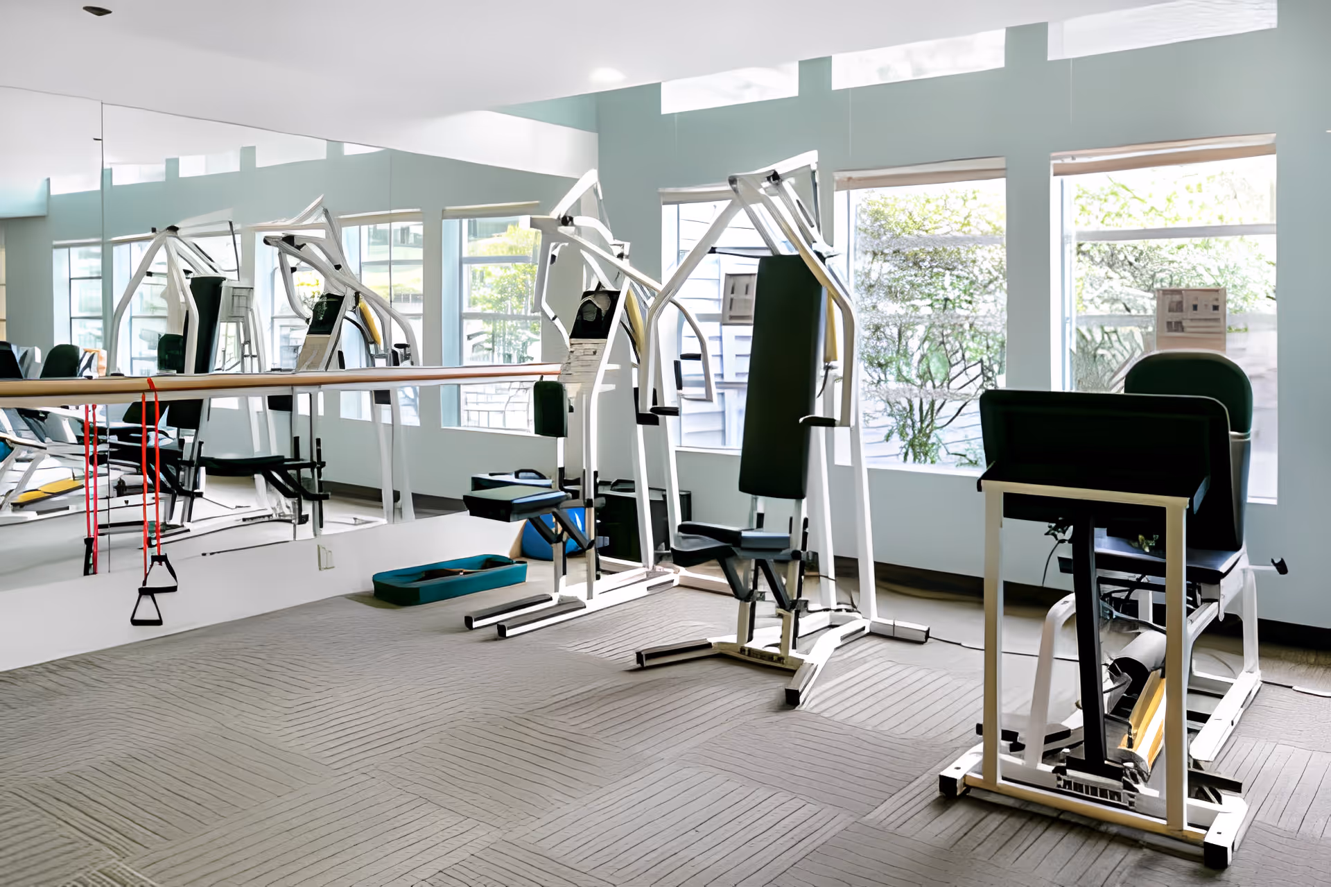 A bright exercise room with large windows letting in natural light. The room contains various gym equipment including a seated weight machine, a leg press machine, resistance bands hanging on a mirrored wall, and other workout accessories on the floor. The walls are painted light blue and the floor is carpeted.