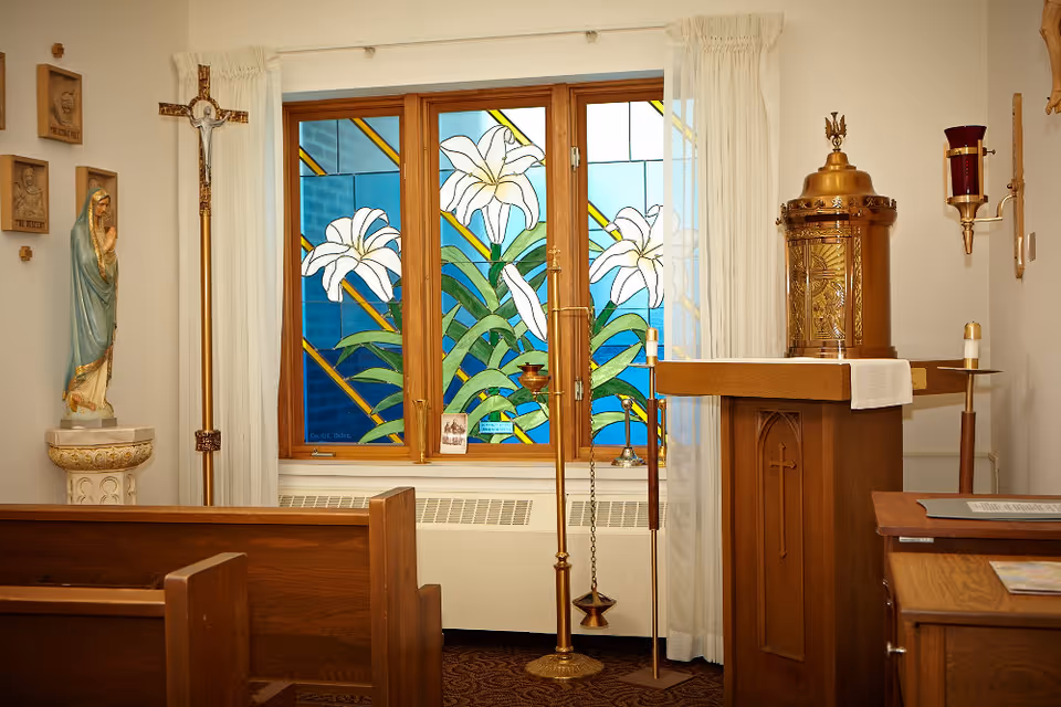 Interior view of a small chapel room with wooden pews, a lectern with a white cloth, a brass tabernacle, a crucifix, a statue of the Virgin Mary, and a stained glass window featuring white lilies and green leaves.