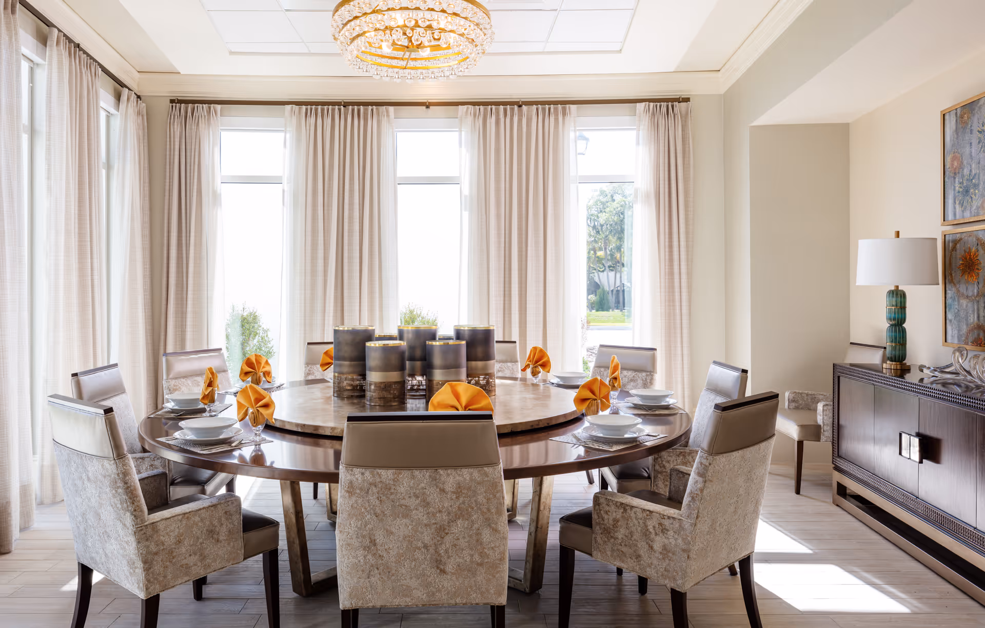 A bright dining room with a large round wooden table set for eight people. Each place setting includes a white bowl, plate, and silverware with an orange folded napkin. The room features tall windows with sheer curtains, a decorative chandelier, and a sideboard with a lamp and artwork on the wall.