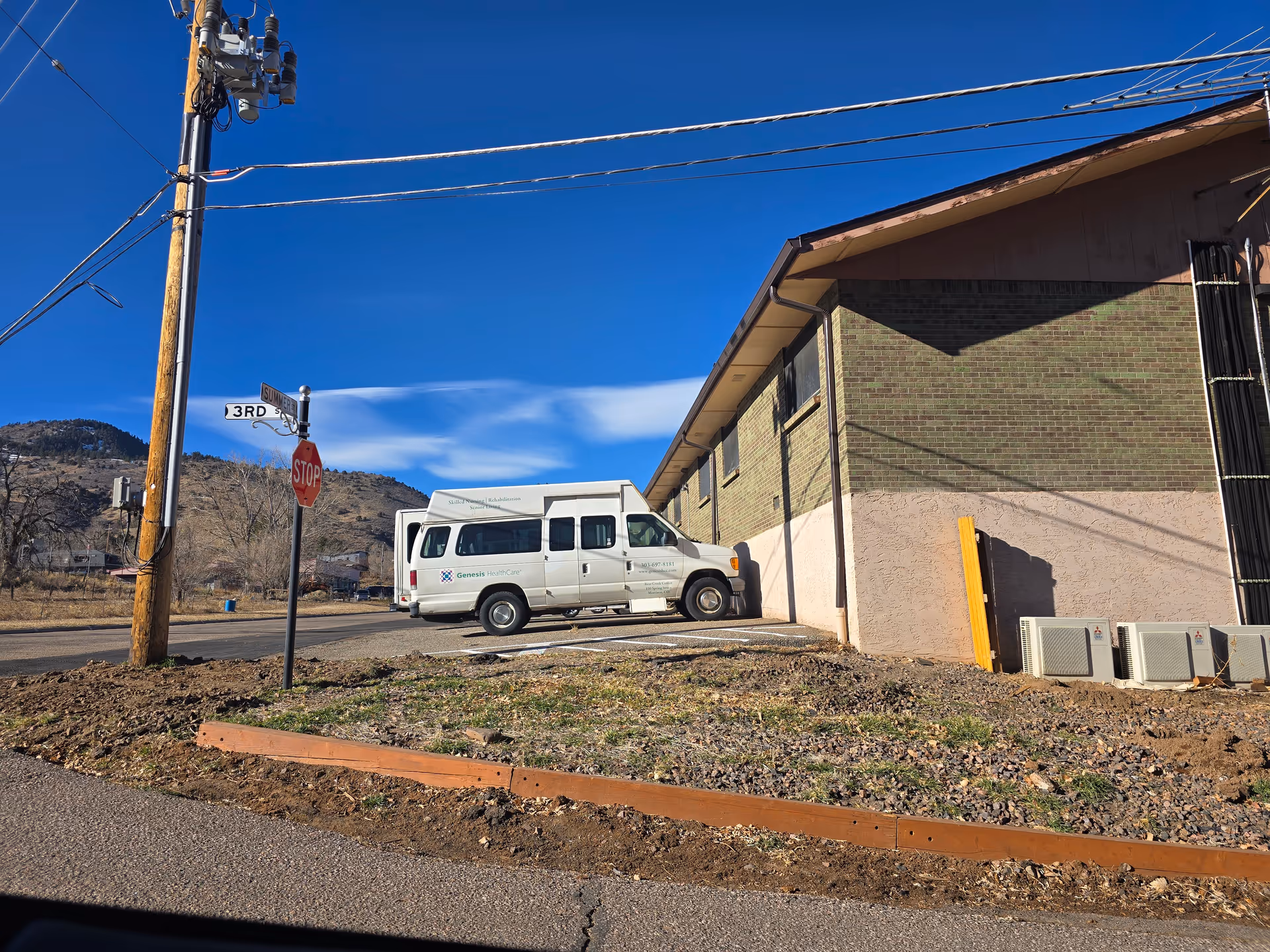 Exterior view of a single-story brick care facility at a street corner with a white shuttle van parked beside it, a stop sign, power pole, and hills in the background.