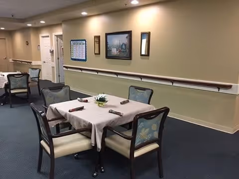 A dining area with a square table covered with a beige tablecloth and four chairs with floral upholstery. The table is set with rolled napkins and a small floral centerpiece. The room has beige walls with framed pictures and a bulletin board, and a handrail runs along the wall. The floor is carpeted in dark blue.