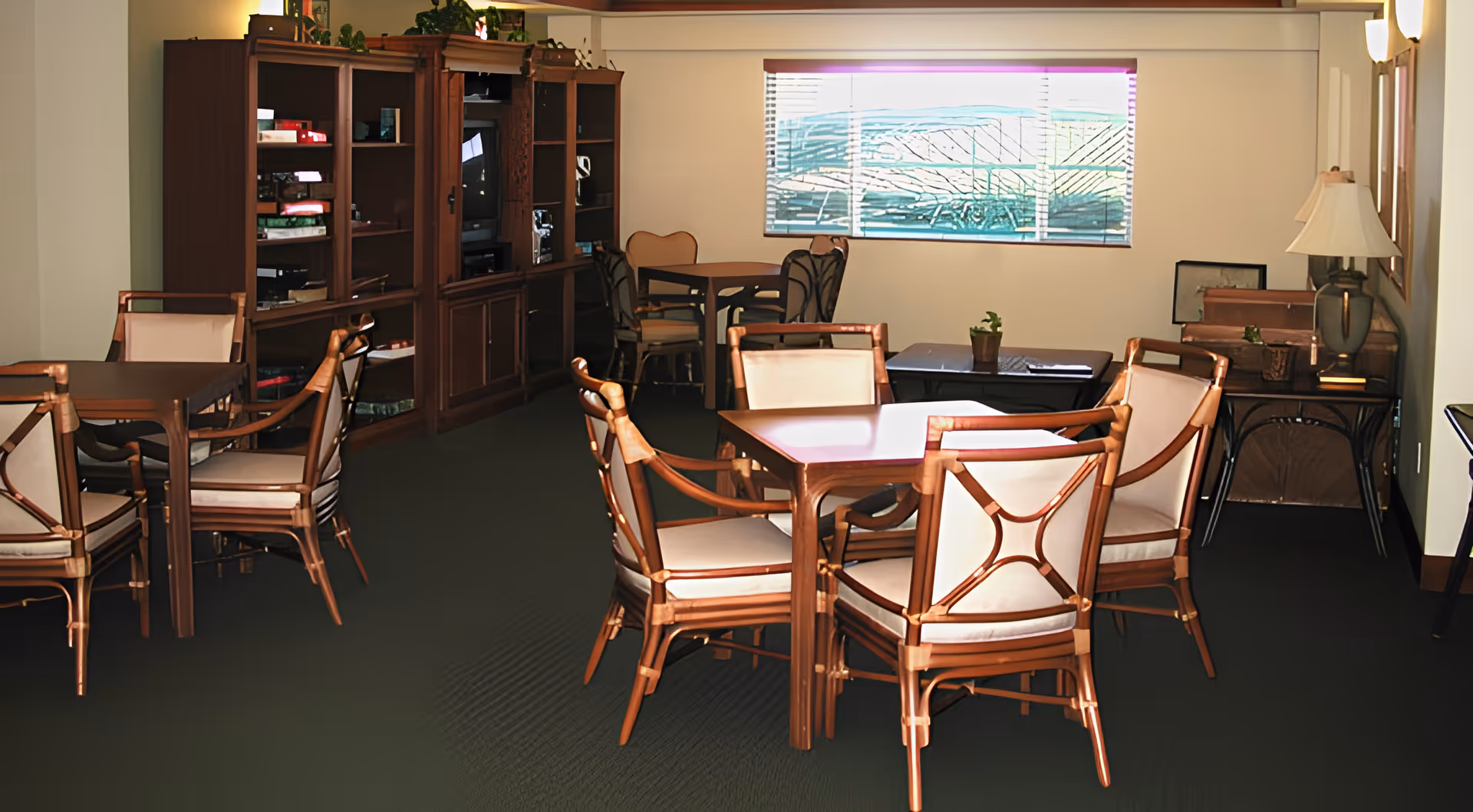 A cozy interior room with several wooden tables and cushioned chairs arranged for seating. There is a large wooden cabinet with shelves and a TV on the left side. A window with blinds lets in natural light, and a table lamp sits on a side table on the right. The room has a warm and inviting atmosphere.