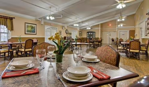A spacious dining room with multiple tables set for meals, featuring plates, bowls, glasses, and red napkins. The room has wooden flooring, ceiling fans with lights, and windows with curtains. A vase with yellow flowers is placed on the table in the foreground.