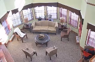 A spacious living room with a beige sofa, four armchairs arranged around a round coffee table, and several windows with wooden blinds letting in natural light. The room has light green walls and a carpeted floor.