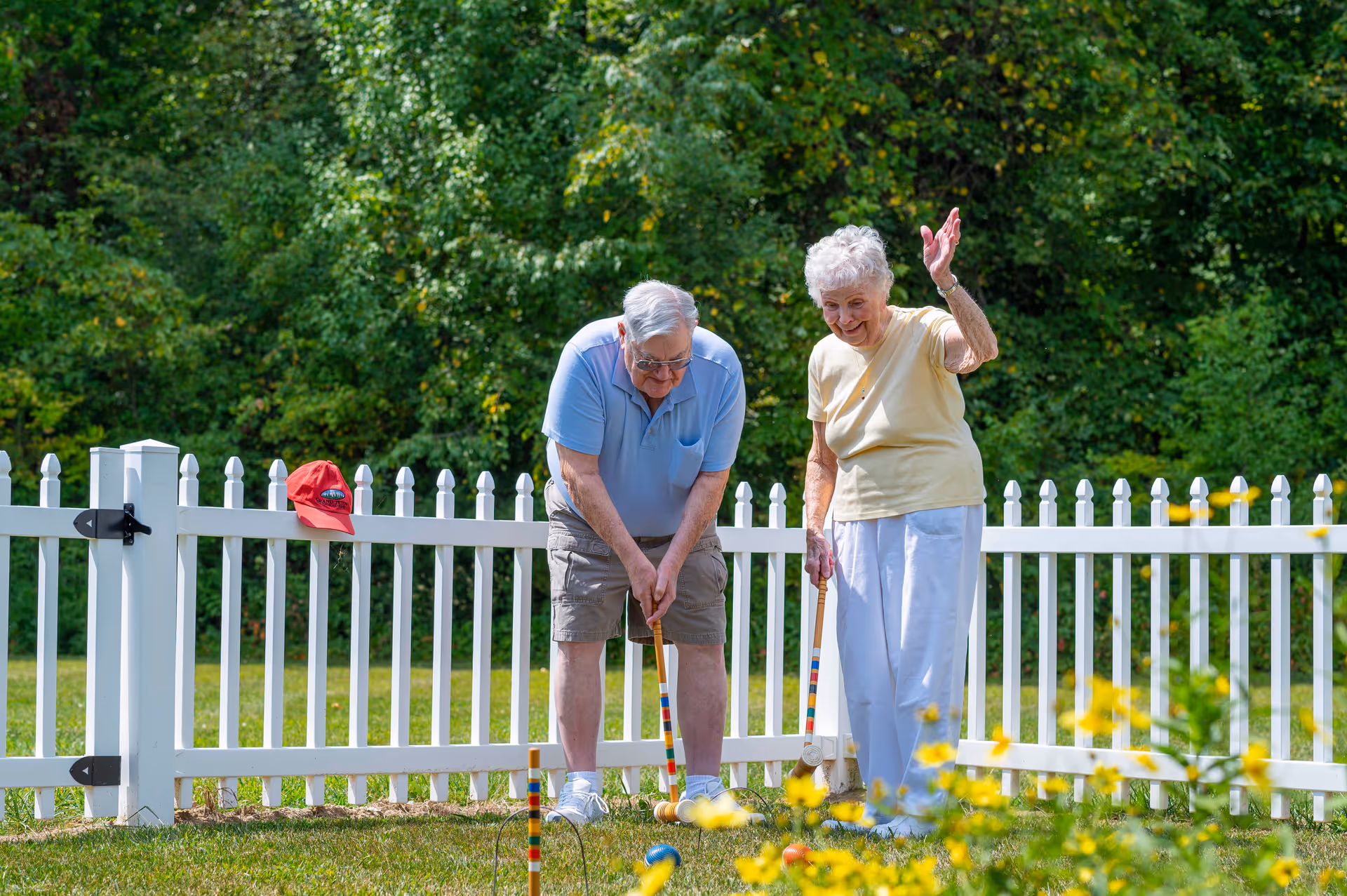 An elderly man and woman playing croquet on a grassy lawn enclosed by a white picket fence. The man is focused on hitting the ball with a mallet, while the woman smiles and waves her hand. There is a red cap hanging on the fence behind them and green trees in the background.