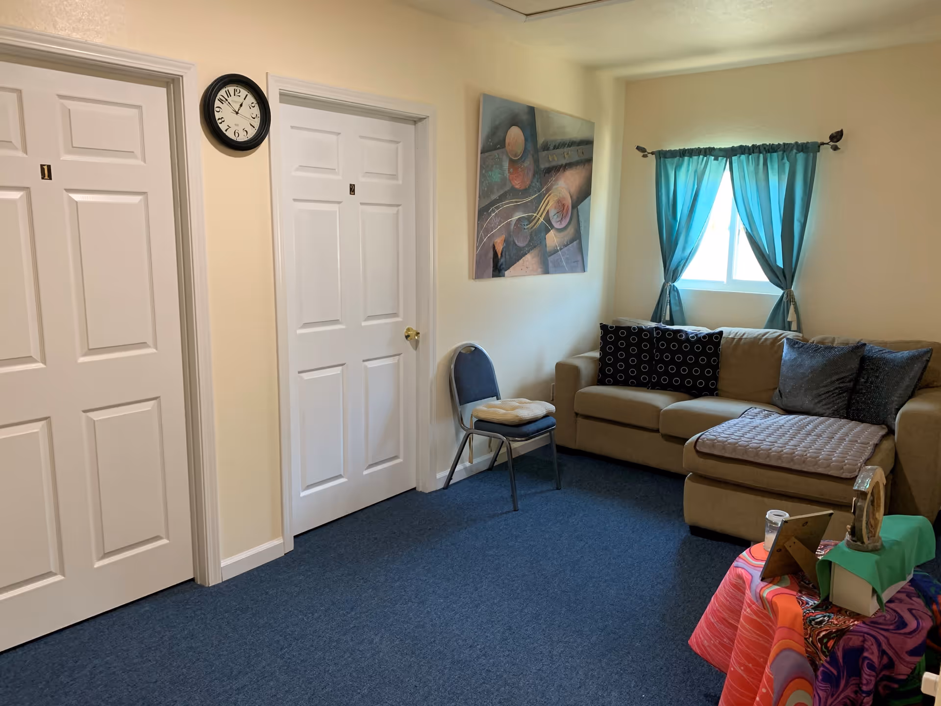 Small lounge with a beige sectional sofa, a chair, two white numbered doors, a wall clock, and a window with teal curtains.