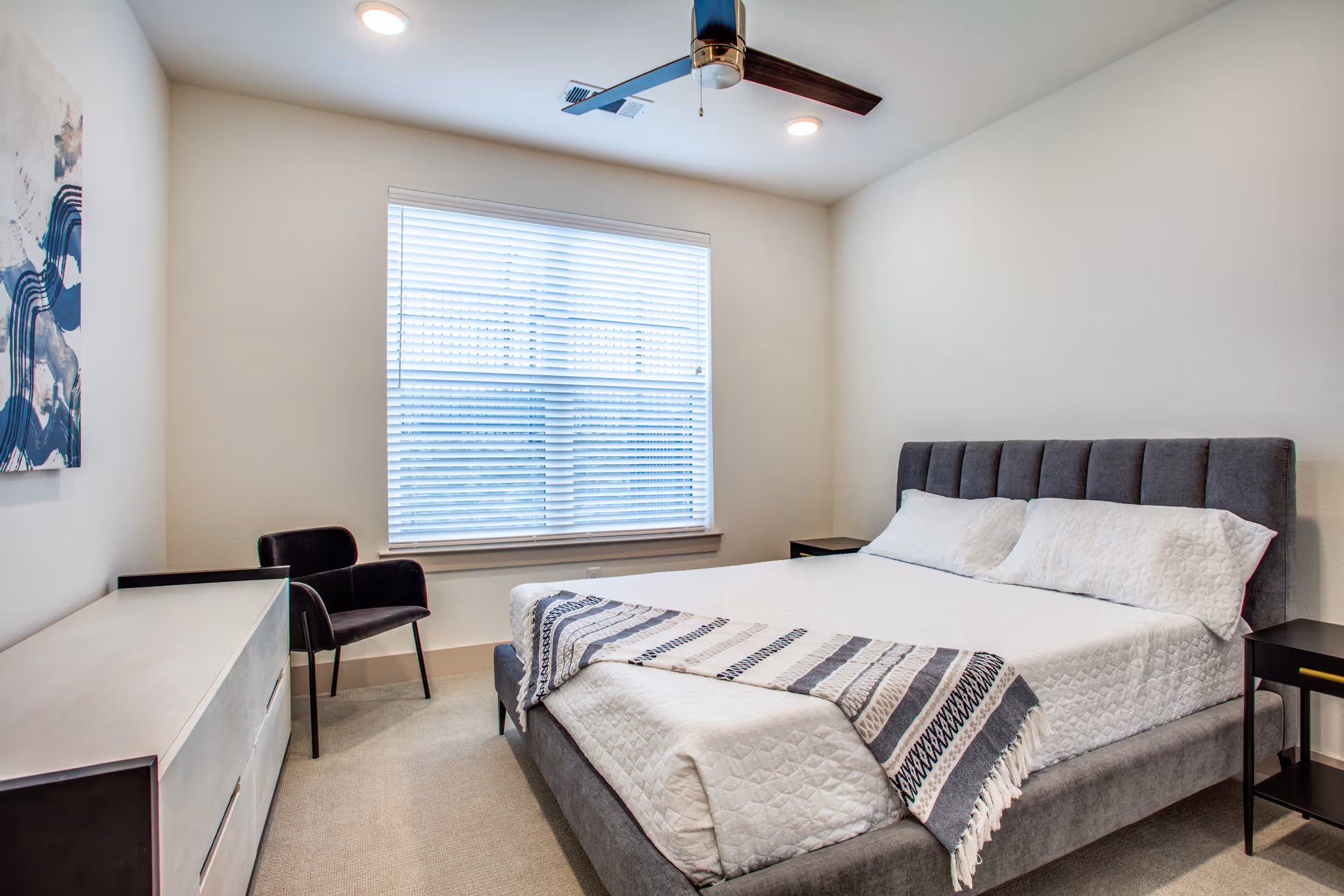 A modern bedroom with a large gray upholstered bed with white bedding and a striped throw blanket. There are two black nightstands on either side of the bed, a black chair near a large window with blinds, a light-colored dresser, and a ceiling fan with lights.