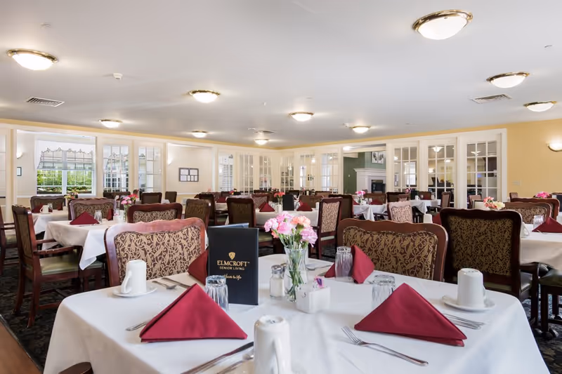 A spacious dining room with multiple tables covered in white tablecloths, each set with burgundy folded napkins, cups, glasses, and silverware. The chairs have patterned upholstery, and there are vases with pink flowers on the tables. The room has large windows and glass-paneled doors, with warm lighting from ceiling fixtures.