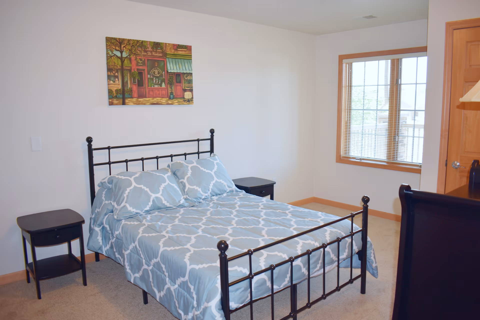 A bedroom with a black metal bed frame and a bed made with light blue and white patterned bedding. There are two black nightstands on either side of the bed. A window with wooden trim and blinds is on the right wall, and a colorful painting hangs above the bed. The room has beige carpet and white walls.