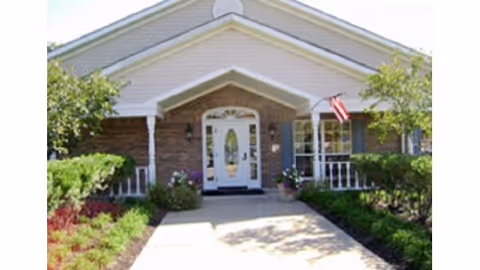 Front entrance of a single-story brick building with a covered porch, white door, American flag and landscaped walkway.