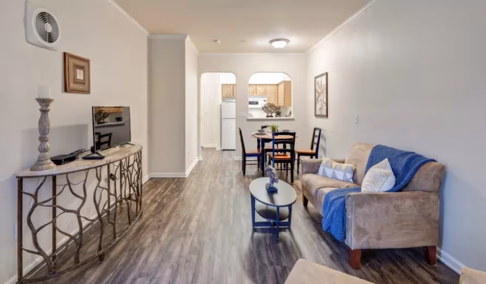 Open-plan living area with a sofa, coffee table, TV console and a small dining table opening to a kitchen.