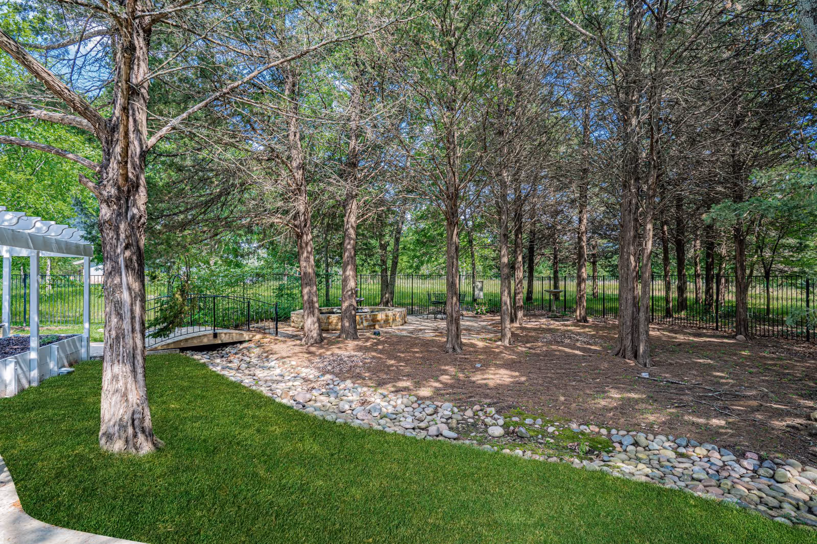 Outdoor garden area with tall trees, a small stone-lined dry creek bed, green grass, a white pergola on the left, and a black metal fence in the background.