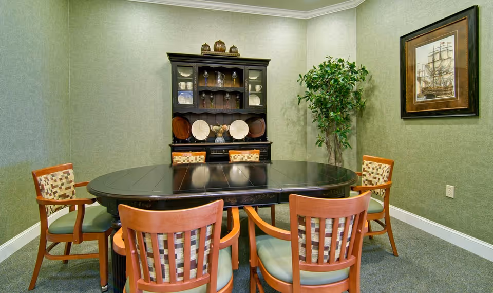 A dining room with a dark wooden oval table surrounded by six wooden chairs with patterned cushions. Against the back wall is a dark wooden hutch displaying plates, glasses, and decorative items. A framed picture of a ship hangs on the right wall, and a tall green plant is placed in the corner.