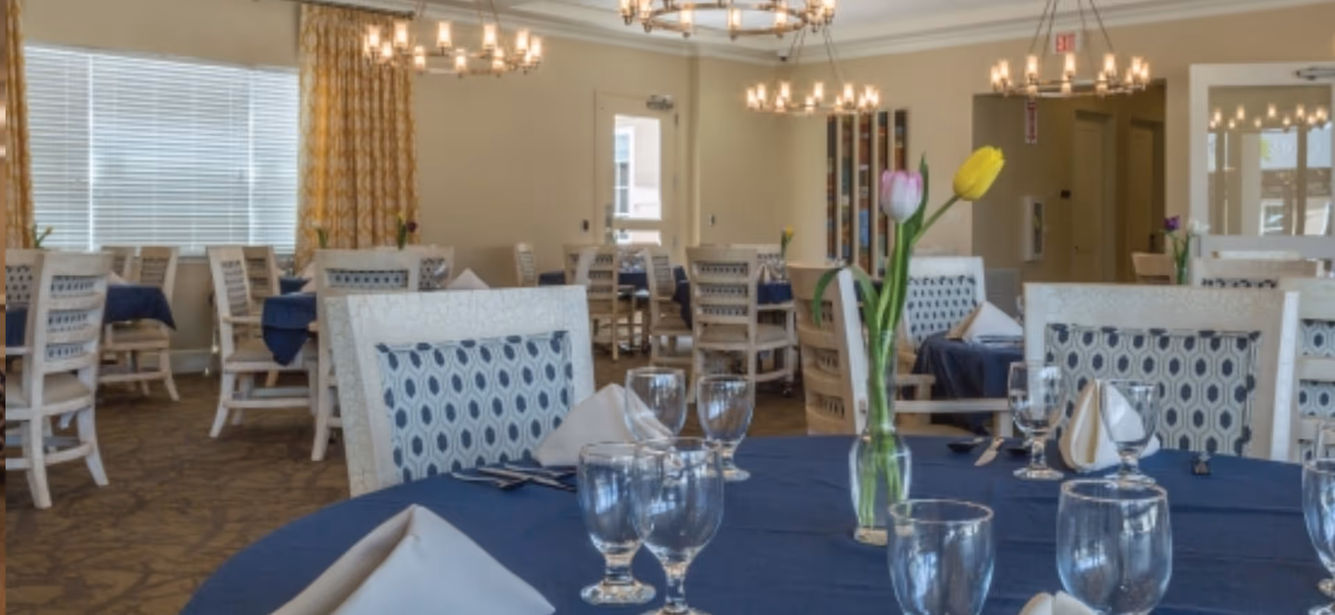 A dining room with multiple tables covered in blue tablecloths and set with glassware, napkins, and silverware. Each table has a small vase with colorful tulips. The room has beige walls, patterned carpet, large windows with yellow curtains, and several chandeliers hanging from the ceiling.