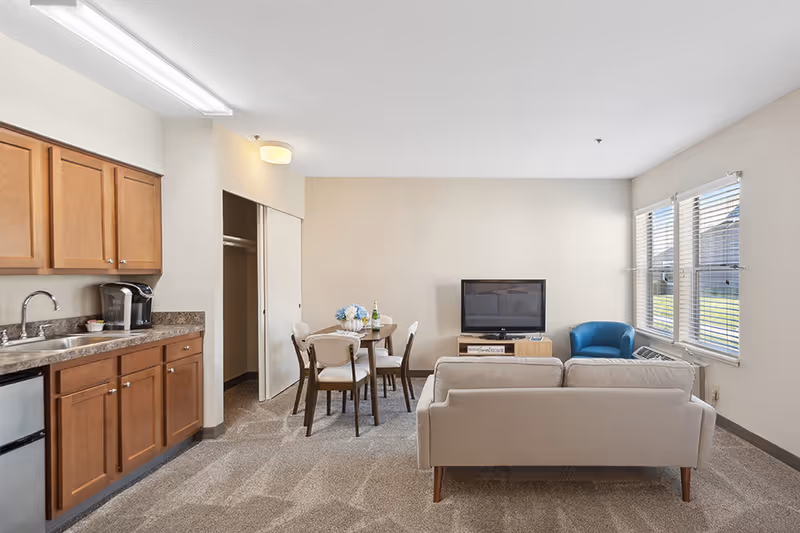 A bright and clean living area in a senior living facility featuring a small kitchen with wooden cabinets and a sink on the left, a round dining table with four chairs in the center, a beige sofa facing a flat-screen TV on a wooden stand, and a blue armchair near two large windows with blinds letting in natural light.