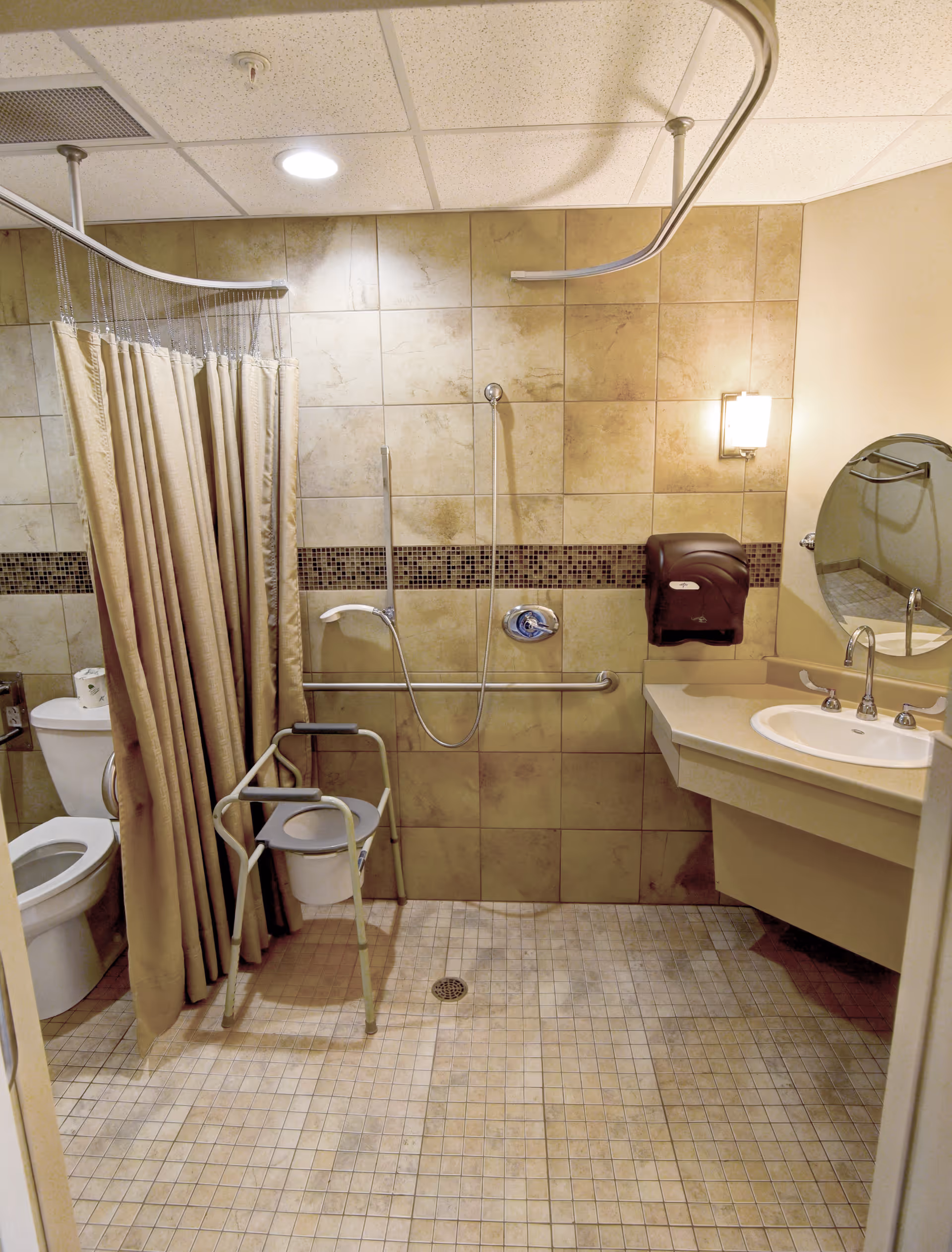 Accessible bathroom with tiled walls and floor, shower curtain and rail, grab bars, shower chair, toilet, sink, and wall-mounted dispenser.