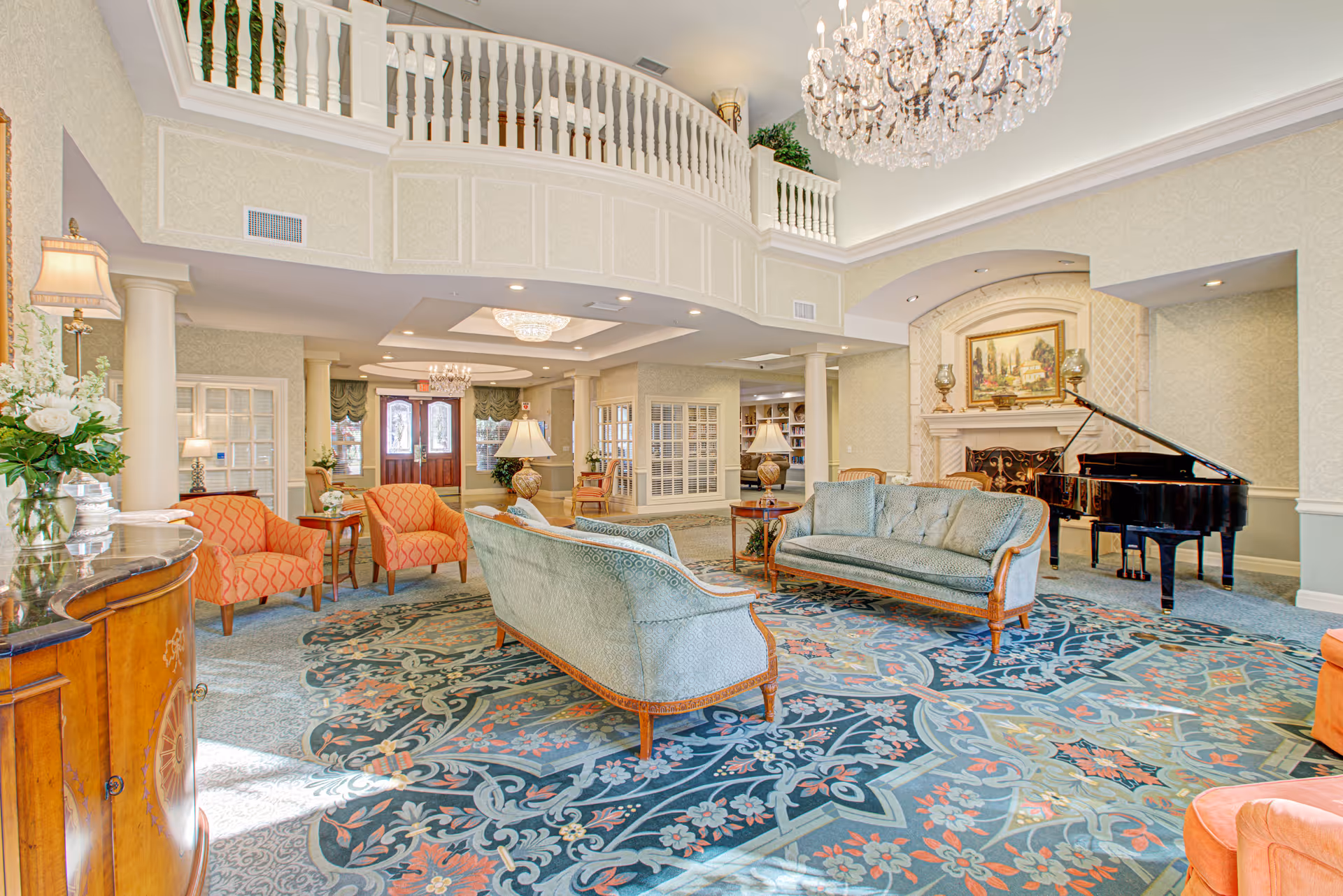 A spacious and elegant living room area in a senior living facility featuring two light blue upholstered sofas, two orange armchairs, a grand piano near a fireplace with a painting above it, a large decorative rug with floral patterns, and a crystal chandelier hanging from the ceiling. The room has a balcony with white railings overlooking the space and several lamps providing warm lighting.