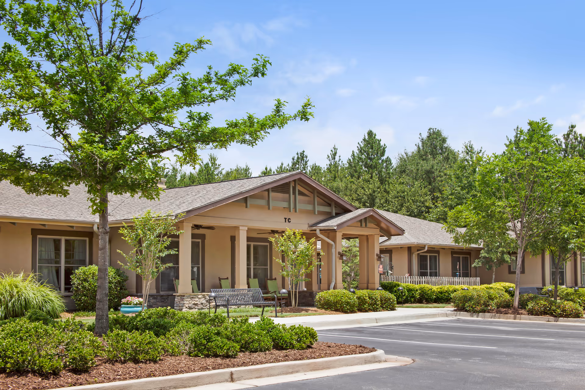 Exterior view of a single-story senior living facility building with a covered porch, green chairs, and benches surrounded by well-maintained landscaping including trees, bushes, and mulch under a clear blue sky.