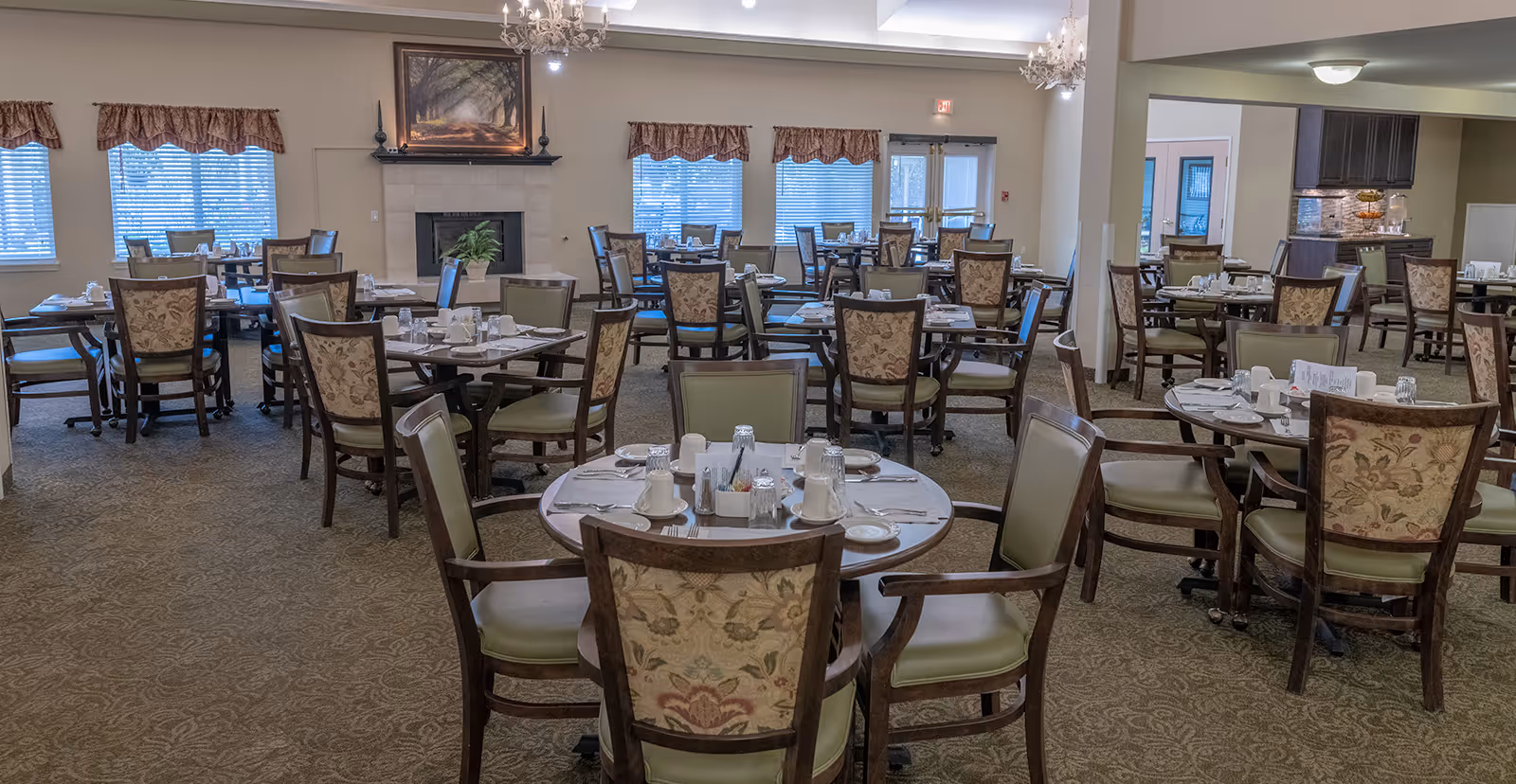 A spacious dining room in a senior living community with multiple round tables set with white cups, plates, and silverware. The room features patterned carpet, cushioned chairs with floral upholstery, large windows with valances, a fireplace with a framed picture above it, and chandeliers hanging from the ceiling.
