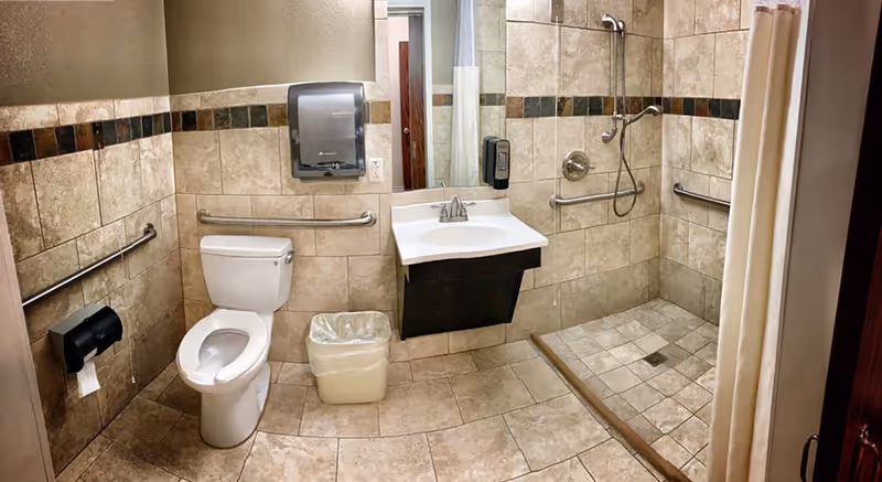 Tiled accessible bathroom with a toilet, wall-mounted sink, paper dispenser, grab bars, and a roll-in shower with a curtain.