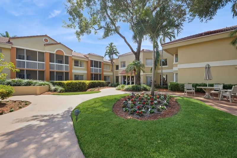 Outdoor courtyard area of a senior living facility with a curved walkway, green grass, a flower bed with red and white flowers, palm trees, and beige two-story buildings with screened balconies and windows under a clear blue sky.