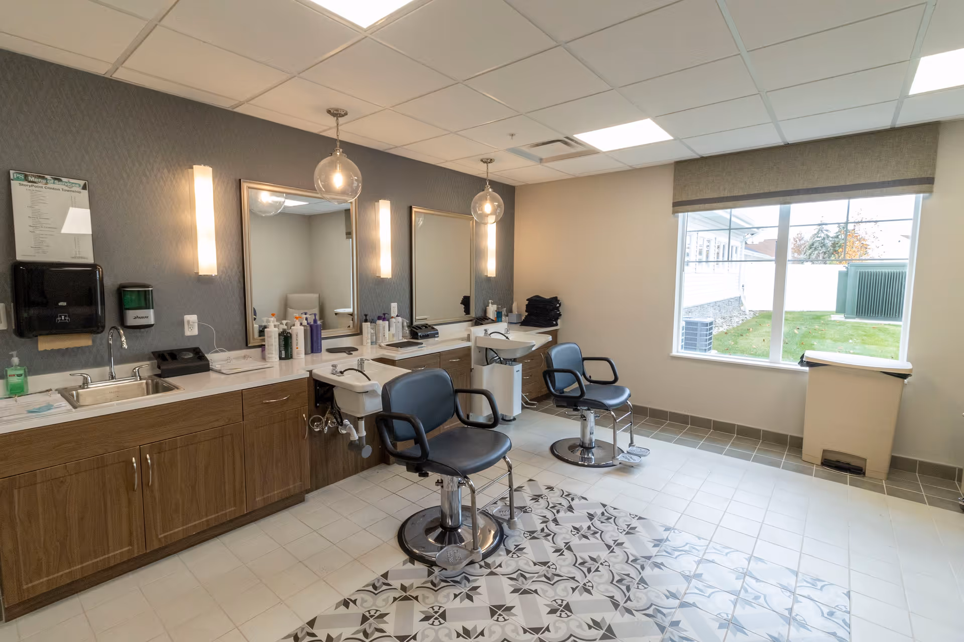 Interior view of a hair salon area in a senior living facility with two black salon chairs in front of two sinks and mirrors. The room has a tiled floor with a patterned section, wooden cabinets, various hair care products on the counter, and a large window showing an outdoor grassy area.