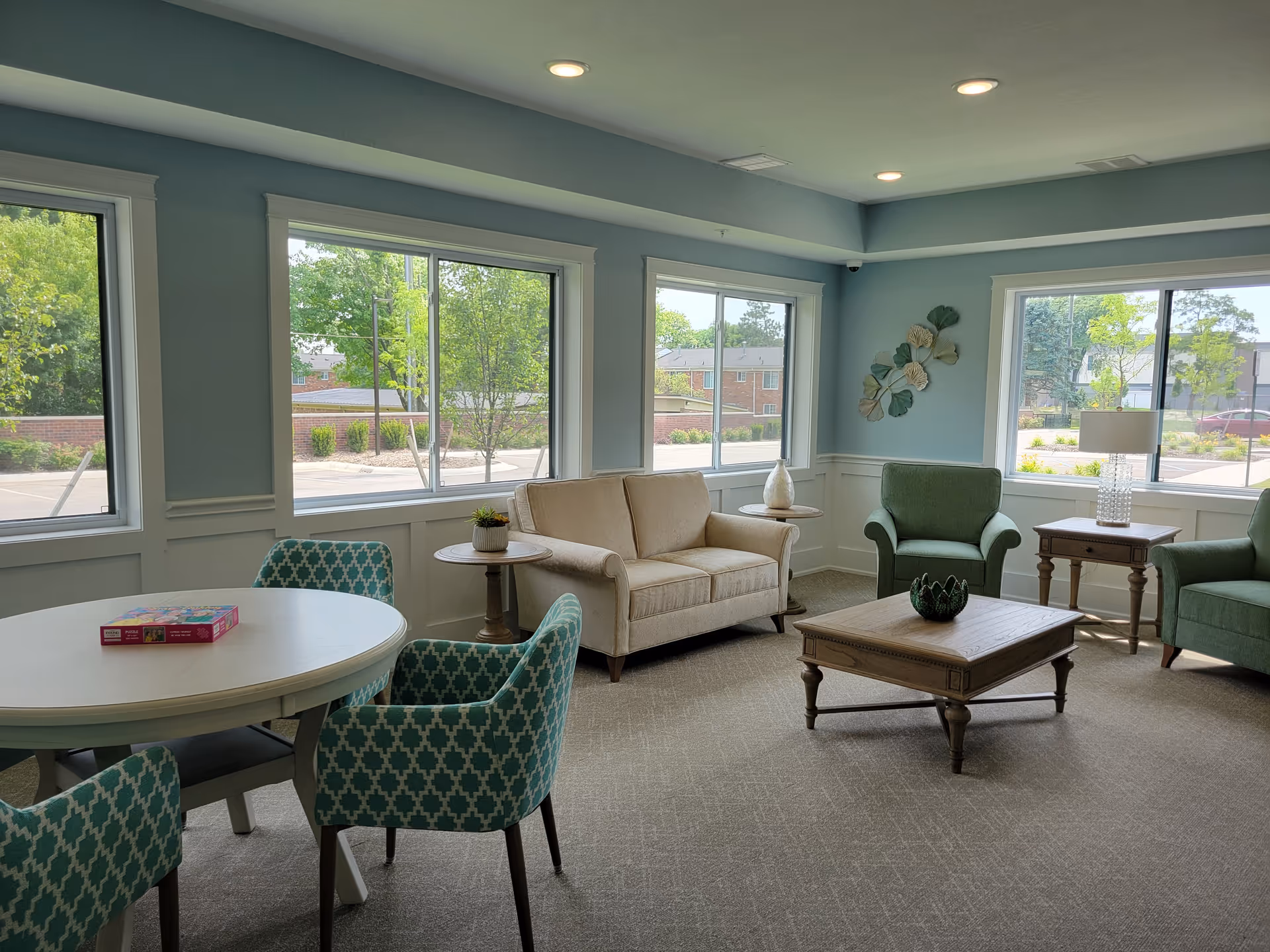 A bright and cozy living room area with large windows showing a view of trees and a parking lot outside. The room features a beige loveseat, two green armchairs, a wooden coffee table with a decorative item, and two side tables with a lamp and a small plant. There is also a round white table with four blue patterned chairs and a board game box on top. The walls are painted light blue with white wainscoting, and the ceiling has recessed lighting.