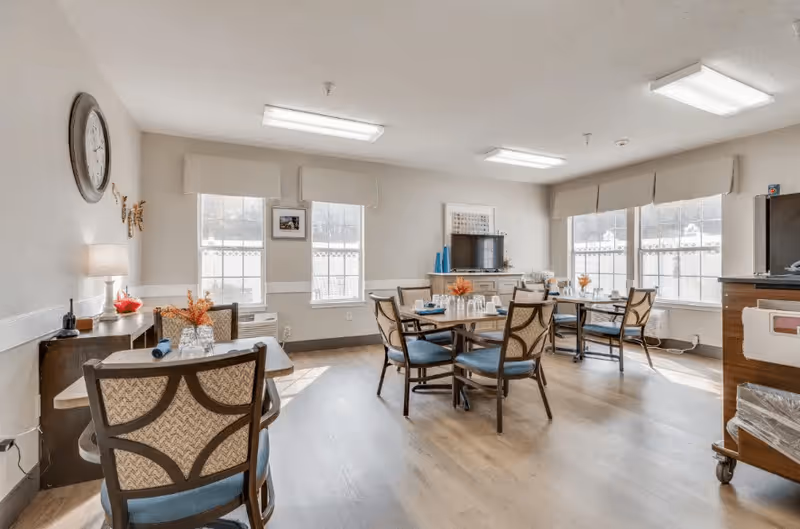 A bright dining room with several tables and chairs arranged neatly. The room has large windows letting in natural light, a wall clock, and a TV on a cabinet. Tables are set with glasses, napkins, and small flower arrangements.