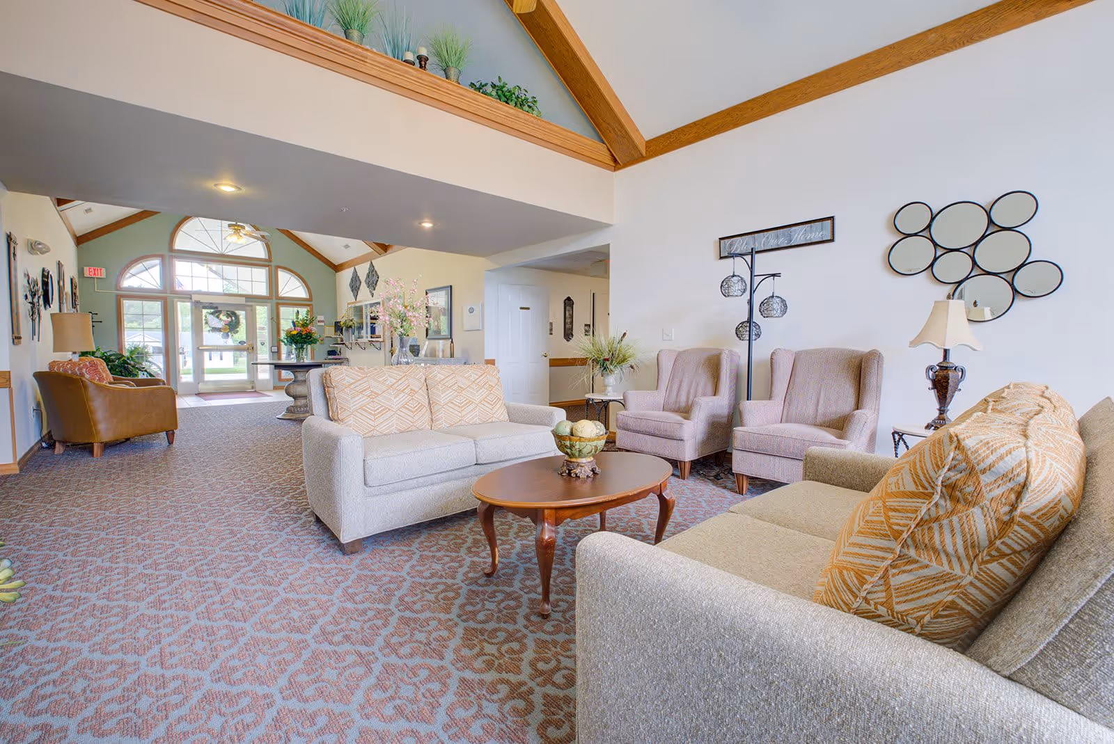 A bright and spacious living room area in a senior living facility with beige sofas, patterned cushions, two armchairs, a wooden coffee table with decorative items, and a lamp on a side table. The room has high ceilings with wooden beams, a patterned carpet, and large windows near the entrance letting in natural light. Decorative wall art and plants add a welcoming touch.