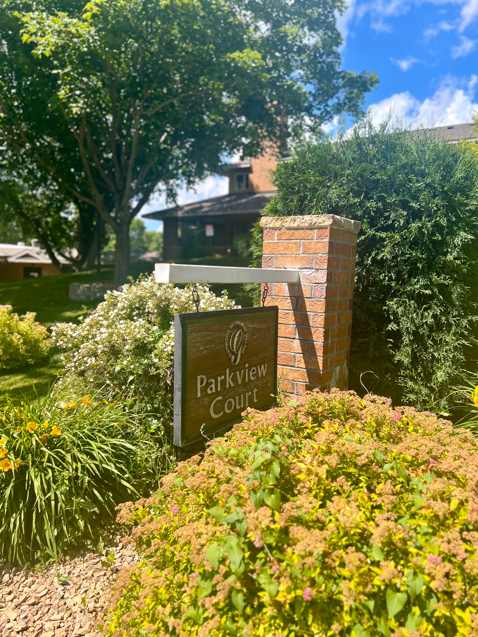 A wooden sign hanging from a white horizontal beam attached to a brick pillar, surrounded by lush green bushes and flowering plants under a bright blue sky with some clouds. The sign reads 'Parkview Court'.