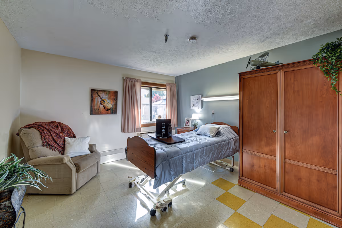 A well-lit room with a hospital-style bed covered with a gray blanket and pillows. Next to the bed is a wooden wardrobe and a small nightstand with a lamp and framed photo. A beige armchair with a red throw blanket and white pillow is positioned near a window with pink curtains. The floor has a checkered pattern with yellow and beige tiles. There is a painting of a guitar on the wall and a decorative airplane model on top of the wardrobe.