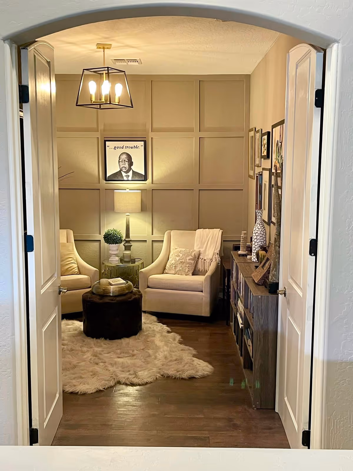 A cozy sitting area viewed through open double doors, featuring two beige armchairs with cushions and a throw blanket, a small round side table with a lamp and a plant, a dark round ottoman on a fluffy white rug, and a wooden bookshelf with decorative items and framed pictures on the wall. A framed portrait with the text '...good trouble.' hangs on the paneled wall behind the chairs.