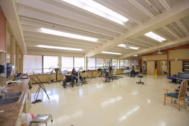 A spacious rehabilitation room with exercise equipment and therapy stations. The room has large windows along one wall letting in natural light, a high ceiling with exposed beams and fluorescent lighting, and a few people engaged in physical therapy activities. There are chairs, a wheelchair, and various exercise machines visible, along with a countertop and sink area on the left side.