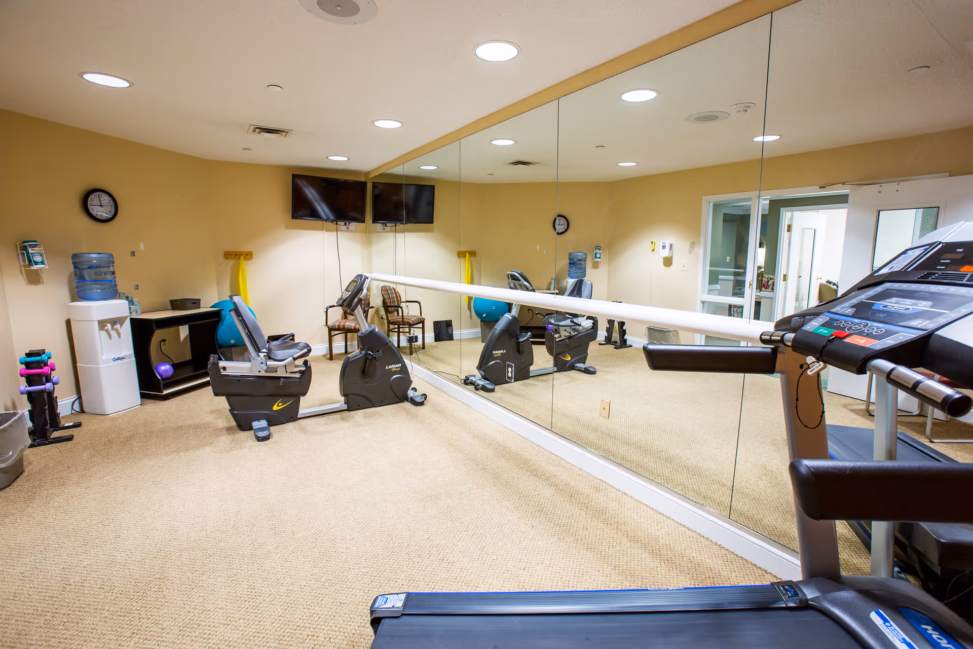 A small fitness room with exercise equipment including a treadmill, two stationary bikes, a set of dumbbells, and a water cooler. The room has beige walls, carpeted floor, a large wall mirror, two wall-mounted TVs, and a clock.