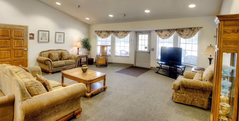 A cozy living room area in a senior living facility with beige upholstered sofas and armchairs arranged around a wooden coffee table. There is a TV on a stand near the windows with floral valances, a wooden rocking chair, a side table with a lamp, and a glass display cabinet with decorative items. The room has carpeted flooring and neutral-colored walls with framed artwork.