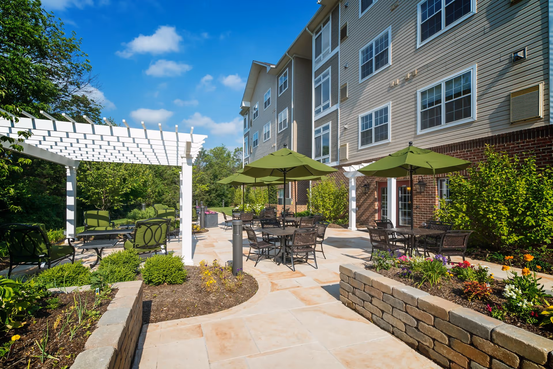 Outdoor patio area at Arbor Terrace Herndon featuring multiple tables with green umbrellas, chairs, a white pergola, and landscaped flower beds with greenery and colorful flowers under a blue sky.