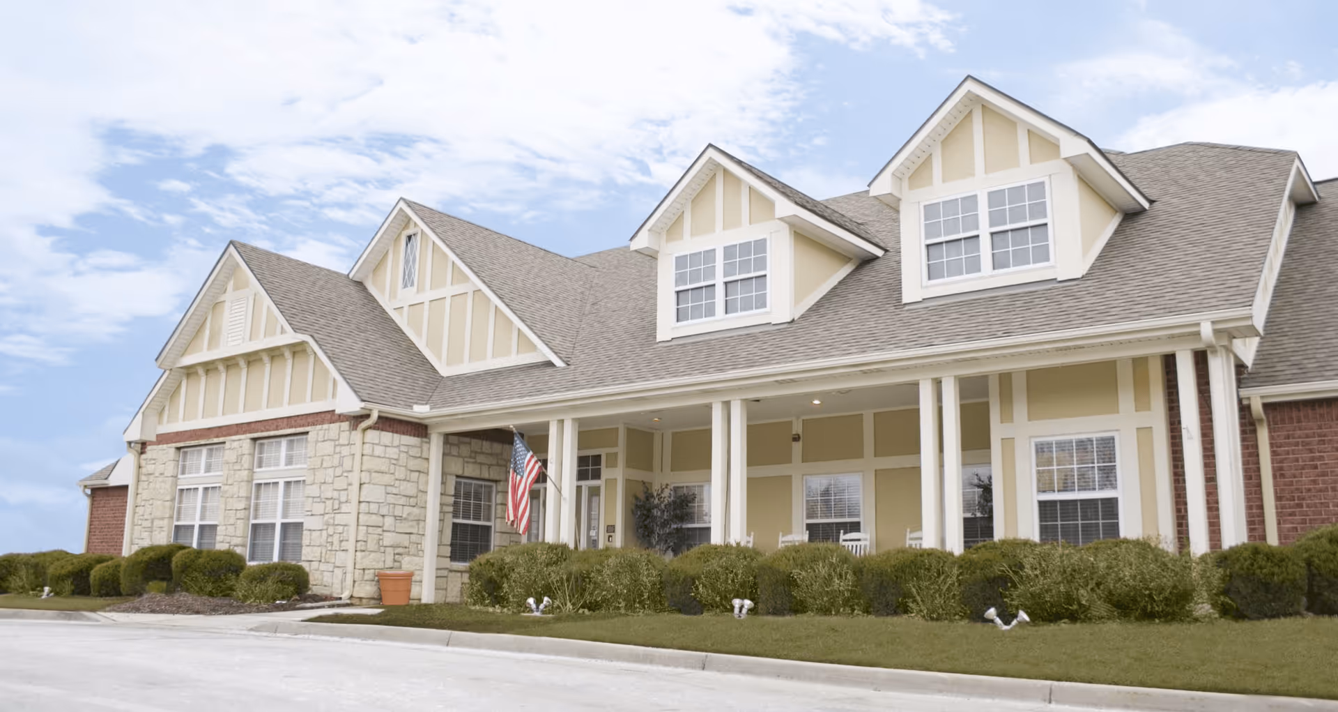 Exterior view of a single-story assisted living facility building with beige and stone facade, multiple windows, a covered porch with white columns, rocking chairs, and an American flag. The building is surrounded by neatly trimmed bushes and a paved driveway.