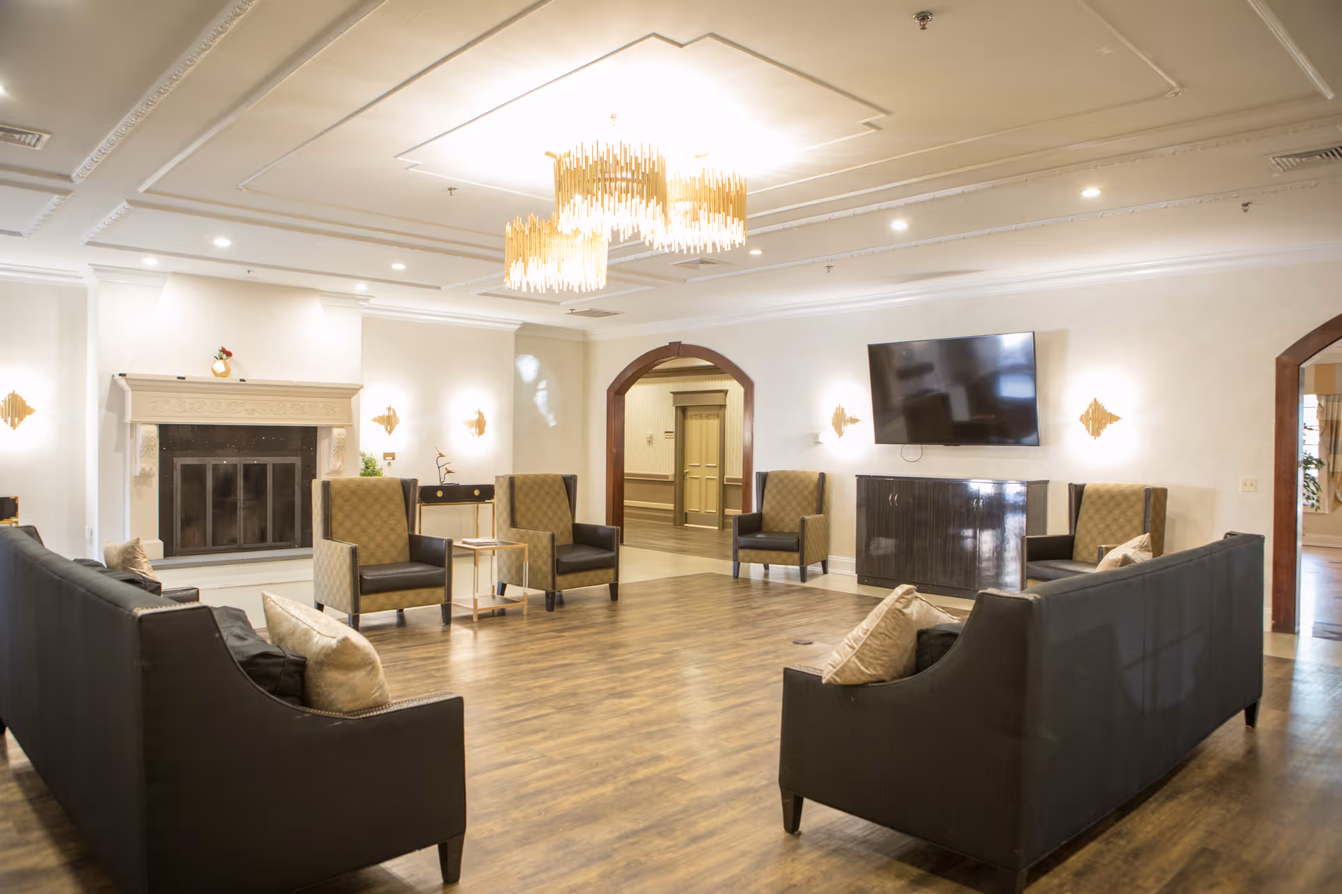A spacious and well-lit living room area in a senior living facility with two black sofas facing each other, four armchairs arranged around the room, a fireplace on the left wall, a flat-screen TV mounted on the right wall, wooden flooring, and a modern chandelier hanging from the ceiling.