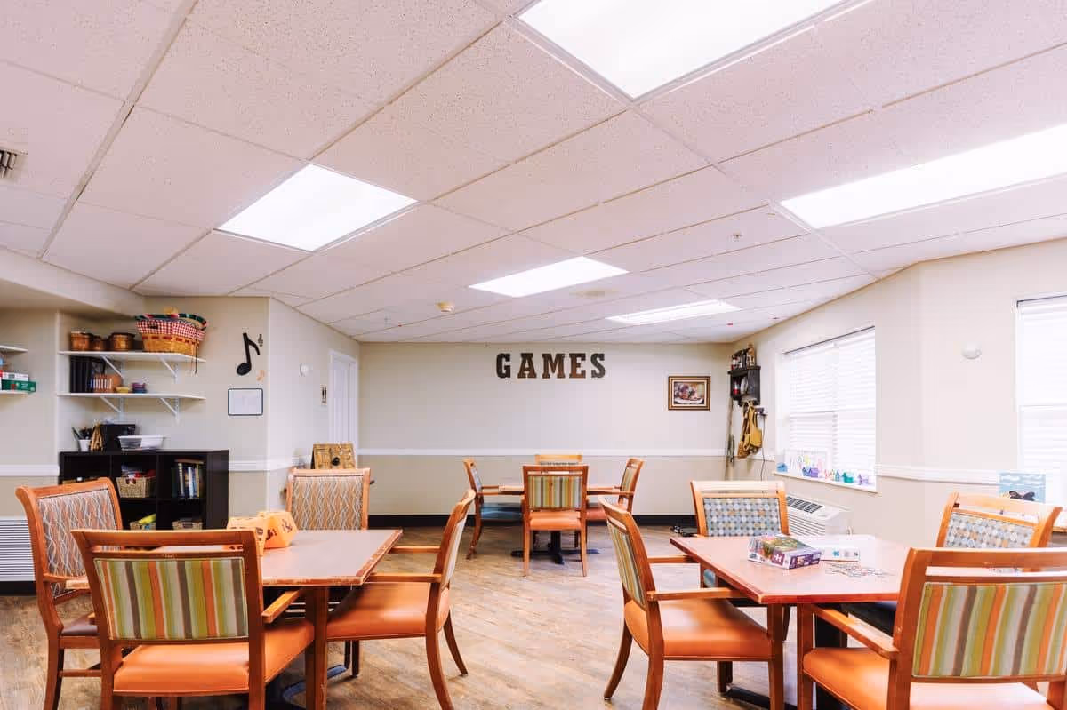 A bright and spacious game room with several wooden tables and chairs arranged for group activities. The walls are light-colored, and the word 'GAMES' is displayed prominently on one wall. Shelves with board games and other items are visible, along with windows letting in natural light.